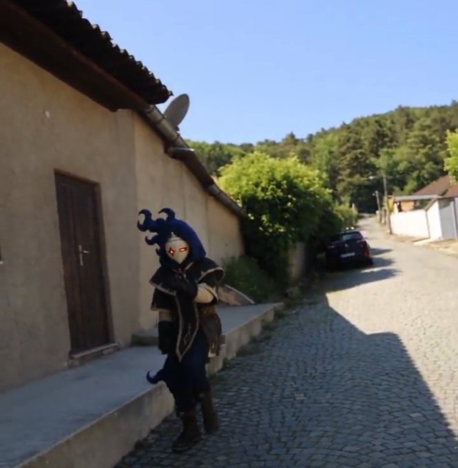 A person in an elaborate dark cosplay costume with tentacle-like hair and a face mask stands on a cobblestone street beside a tan building.