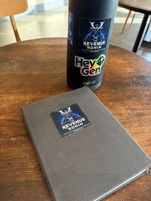 The image features two items, an open notebook with the text "Revenue Ronin" written on its cover, and a black and grey water bottle with a sticker on it that has the words "hey 𝕏 builder". These objects are placed next to each other on a table. The presence of these items in close proximity suggests that someone might be using the notebook as a reference for information about the revenue building process or as a planner, while also having the water bottle nearby for hydration during their work or study sessions.