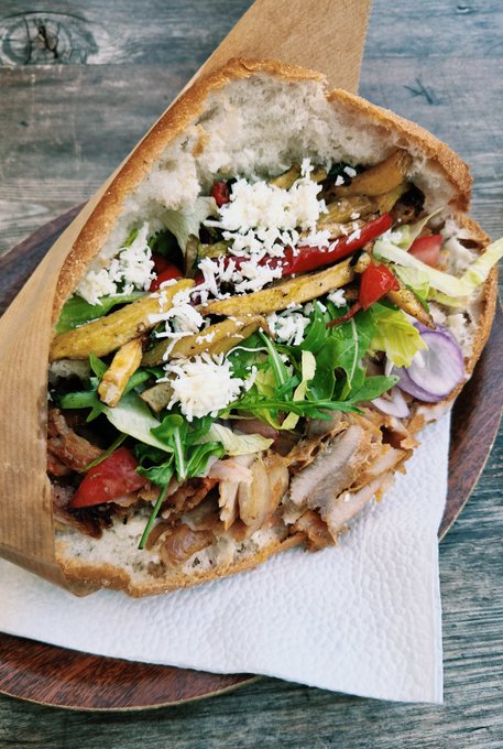 A close-up of a stuffed baguette sandwich on a wooden board shows meat, leafy greens, tomato, onion, yellow vegetable strips, and crumbled white cheese.