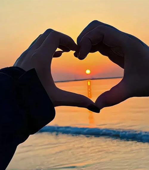 Two hands form a heart shape silhouetted against a sunset over the ocean.