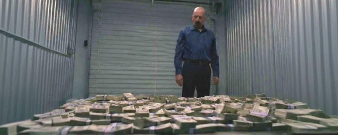 A man in a blue shirt stands behind a large table covered in stacks of US dollar bills inside a room with metal walls.