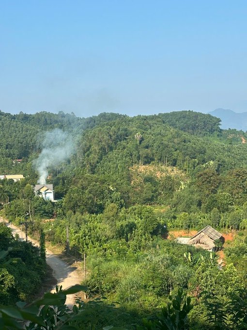 A scenic landscape of lush green hills and trees with a small house and smoke rising in the distance, with mountains visible in the background.