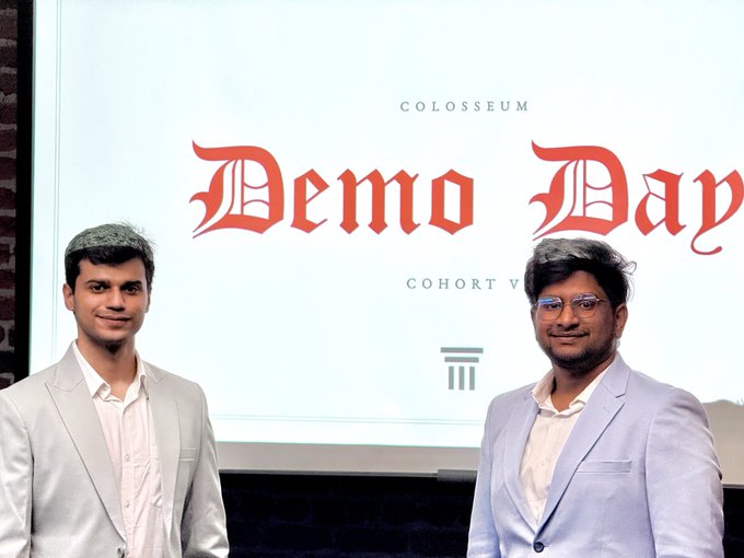 Two men in light-colored blazers standing in front of a presentation screen displaying 'COLOSSEUM Demo Day COHORT V' in red gothic-style text.