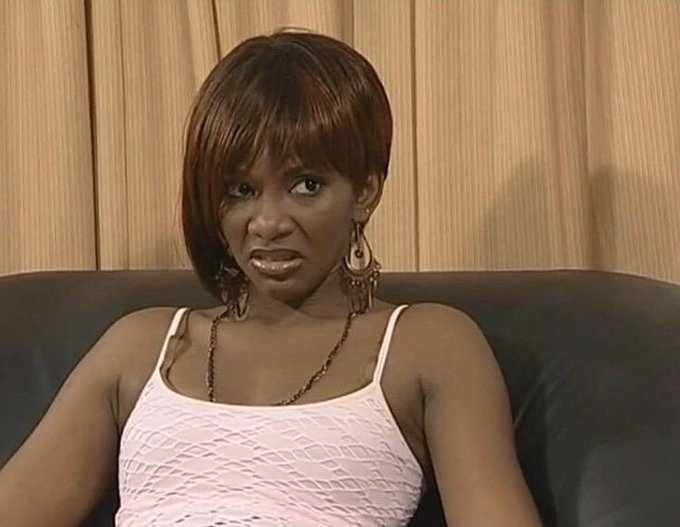 Skeptical-looking woman with short reddish-brown hair and large hoop earrings sitting on a dark couch, wearing a white tank top.