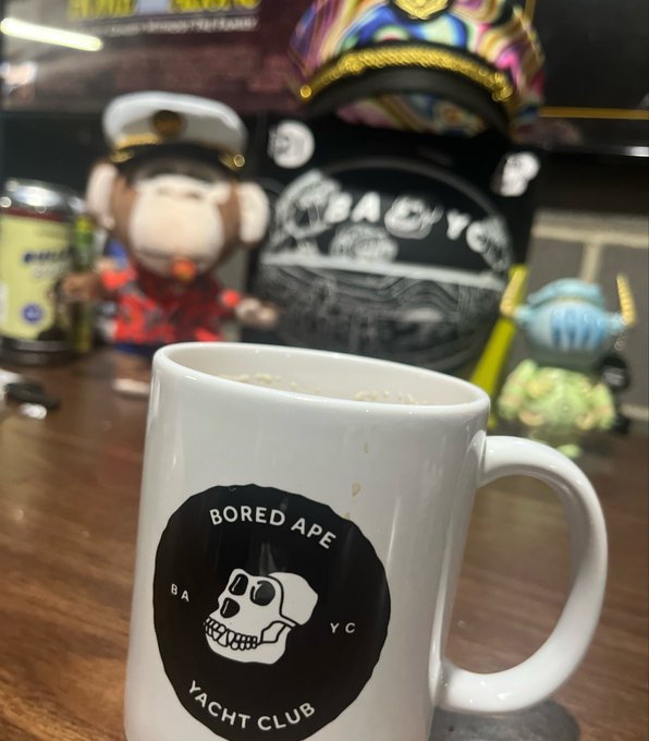 A white Bored Ape Yacht Club (BAYC) mug sits on a wooden table in front of Bored Ape NFT figurines.