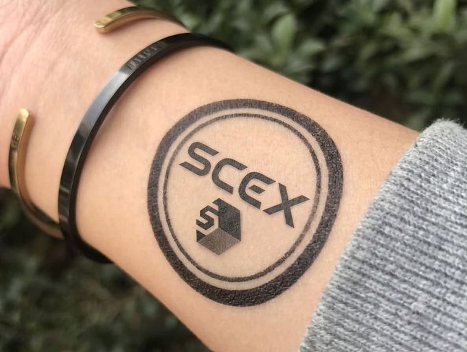 A wrist with a 'SCCEX' tattoo inside a circle, wearing a black bracelet and a gold cuff bracelet.