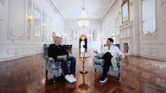 Two men are seated in ornate armchairs facing each other in a grand, palatial room with white walls, gilded mirrors, a chandelier, and a polished parquet floor, with a small table between them.