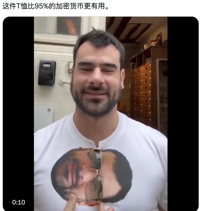 A video still of a smiling man in a white t-shirt with a printed face design, with Chinese text above reading '这件T恤比95%的加密货币更有用。' (This T-shirt is more useful than 95% of cryptocurrencies).