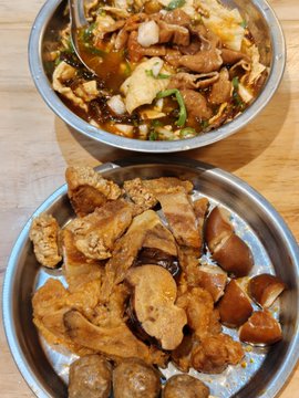 Two metal bowls of food, one containing a dark brothy stew with meat and greens, the other filled with various braised pork parts and offal.