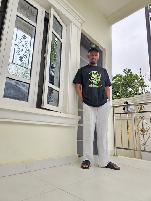 A man wearing a black 'ShipGuild' t-shirt, gray sweatpants, a cap, and sandals on a tiled balcony.