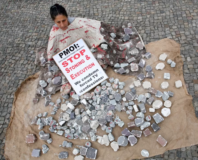 A protest performance featuring a person covered in a blood-stained sheet sitting on a burlap sack surrounded by broken stone fragments on cobblestones, holding a sign that reads 'PMOI: STOP STONING & EXECUTION' with smaller text stating 'We condemn forced TV (confessions)'.