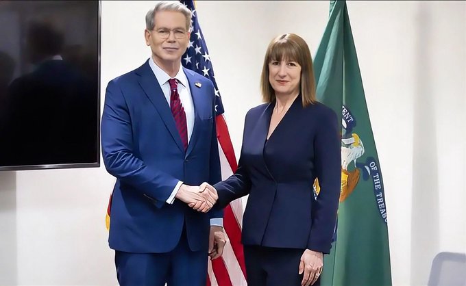 Two officials, a man in a blue suit and red tie and a woman in a navy blazer, shake hands while posing in front of an American flag and a green flag bearing an eagle emblem.