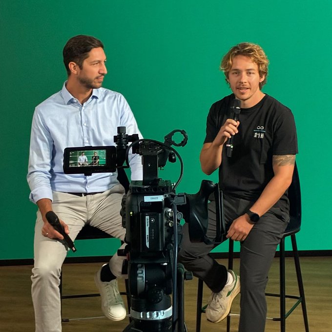 Two men seated in front of a green screen, engaged in a discussion with microphones, with a professional video camera positioned between them recording the session.