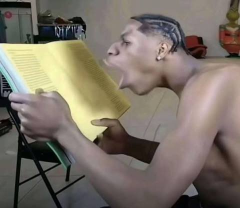 A shirtless man with braided hair intensely reading or yelling from an open book held in his hands in what appears to be a home setting.