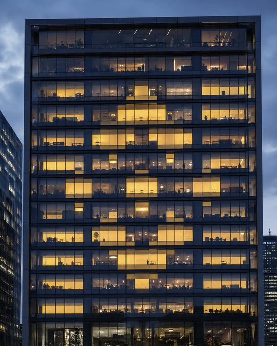 A modern office building stands illuminated against a dusky blue sky, with warm yellow light spilling from its many windows revealing glimpses of interior workspaces, while partial structures of neighboring skyscrapers flank it on either side. The glowing grid-like pattern of lit windows visually echoes the "GN" ticker, suggesting corporate activity and the bright "green" (upward) market performance it represents.