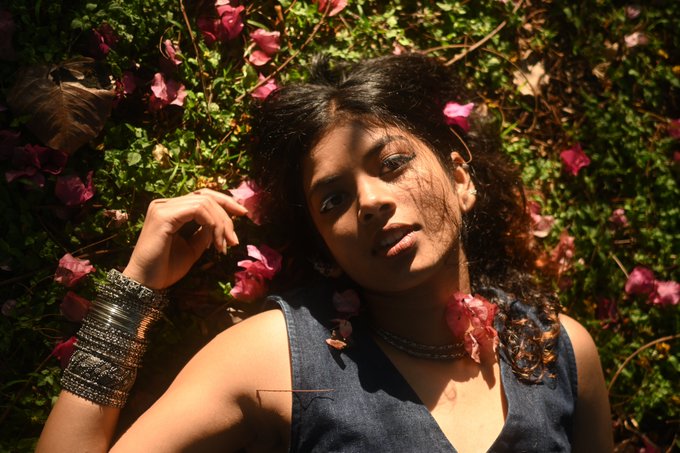 A woman with dark curly hair lies among pink flowers and green foliage, wearing a blue denim top and silver bracelets, with a pink flower resting on her neck.