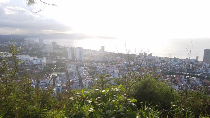 A scenic landscape photograph taken from an elevated vantage point, looking down over a coastal city toward the ocean under a bright, hazy sky, framed by green foliage in the foreground.