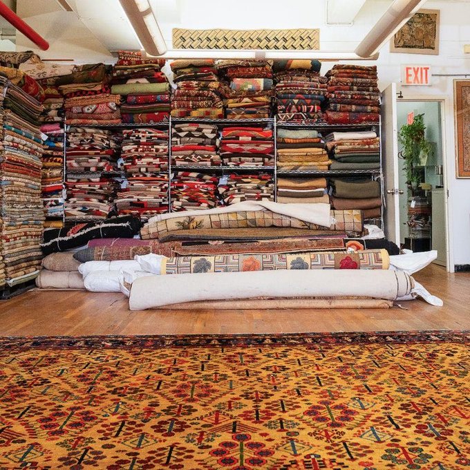 A rug store interior stacked floor-to-ceiling with colorful oriental carpets and rugs displayed on shelves, with several rolled rugs lined up in the foreground. The "EXIT" sign is visible above the door, though the joke in the caption hinges on the "rug" connection to Binance, the cryptocurrency exchange whose name means "coin exchange" — playing on the double meaning of "rug" as both a floor covering and the crypto slang for a sudden price crash ("getting rug-pulled").