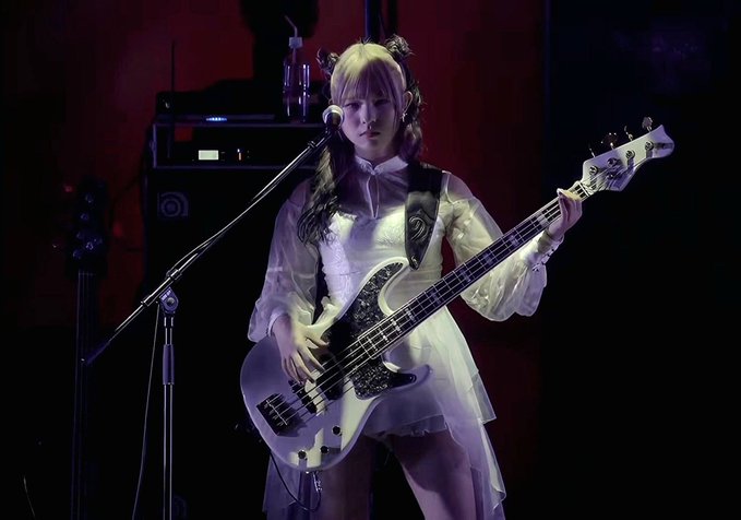 A blonde woman in a white dress plays a bass guitar on stage under purple lighting.