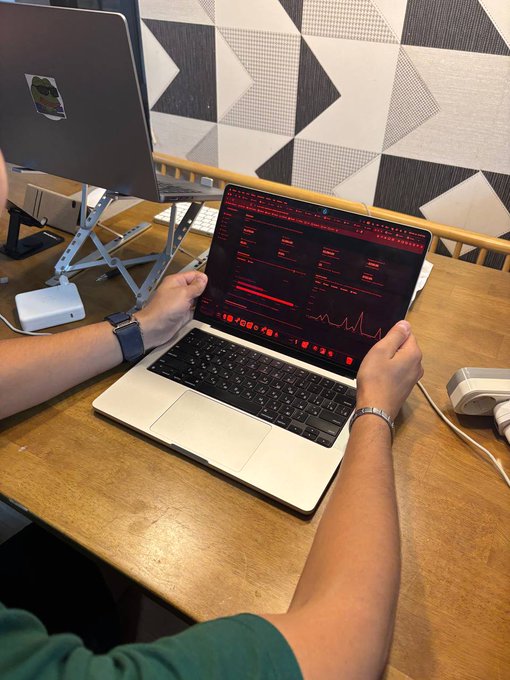 A person wearing a green shirt sits at a wooden desk using a laptop displaying red and black financial charts, with a monitor mounted on a stand in the background.