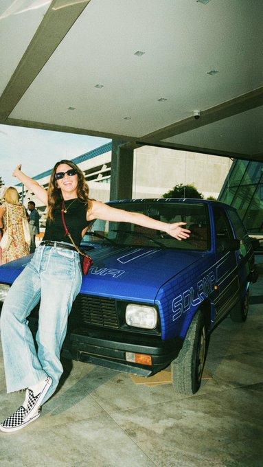 A woman in jeans, sunglasses, and checkered Vans poses on the hood of a blue 'Solar' truck in a film-photo aesthetic.