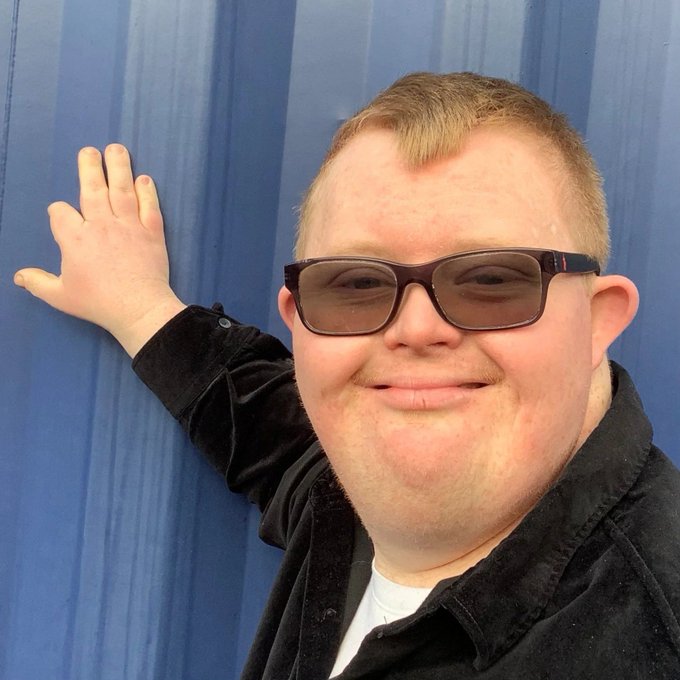 A man with short blonde hair and tinted sunglasses smiles at the camera while leaning against a blue padded wall with one hand raised.