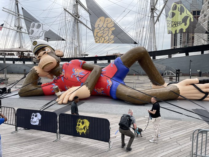 The image features a large inflatable monkey statue, possibly from the movie "A Walk on Water", lying on its back with arms and legs spread out. The monkey is surrounded by several people who are standing around it or walking near it. In addition to the monkey, there are also two boats visible in the background - one of which has a skull design painted on it. The scene appears to be at an event related to "A Walk on Water", as indicated by the presence of the inflatable monkey and the people gathered around it.