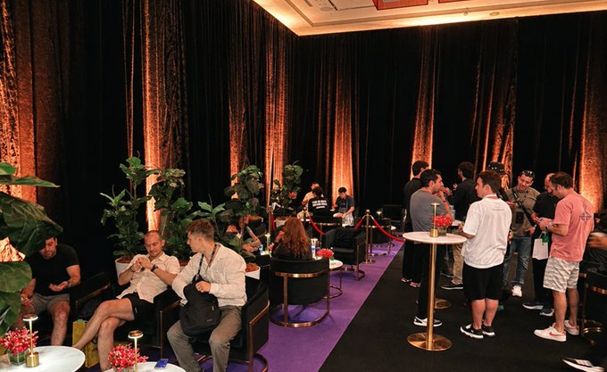 An indoor event space with black curtains and warm lighting shows attendees mingling around cocktail tables, some seated and others standing in conversation near plants and bar stools.