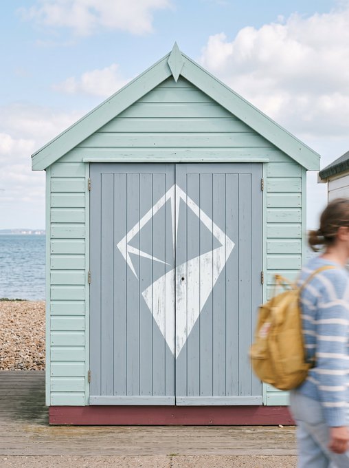 A light blue wooden beach hut features a large white Ethereum logo painted on its double doors, with a person carrying a yellow backpack walking past.