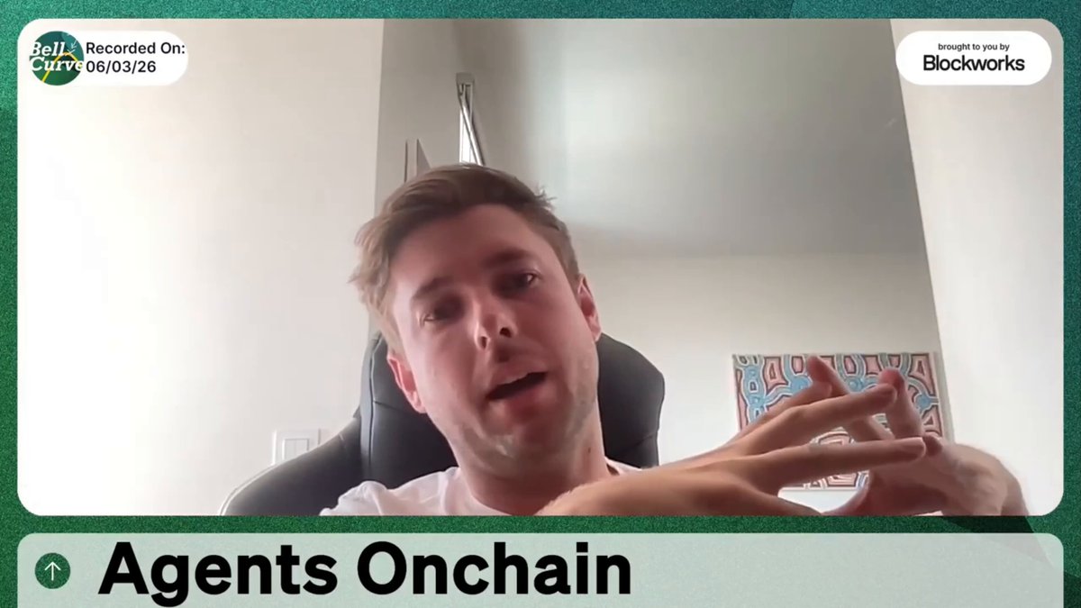 The image shows a man sitting in an office chair, wearing a white shirt with black text that says "Agents Onchain" written below him. The background of the image features a green wall with a painting hanging above it.