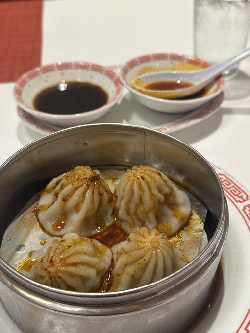 A metal steamer basket holds four soup dumplings (xiaolongbao) drizzled with sauce, alongside small dishes of soy sauce and vinegar on a table with red-patterned plates.