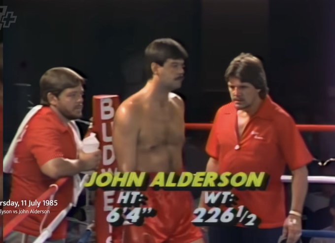 A muscular man stands in a boxing ring flanked by two men in red shirts, with on-screen graphics displaying "JOHN ALDERSON," HT: 6'4", WT: 226½, dated Thursday, 11 July 1985. The caption jokes about "crypto," likely a playful reference to the man's notably chiseled, "sculpted" physique resembling cryptocurrency icons or just the absurd randomness of pairing a vintage boxing broadcast with a modern crypto joke.