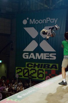 A skateboarder performs an aerial trick above a ramp at the MoonPay X Games Chiba 2026 event, with a person in a green shirt watching from the side. The caption "thinking about ramps" ties into the image by highlighting the prominent skateboard ramp visible in the foreground.