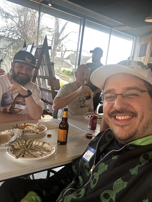 A selfie of three men smiling at a restaurant or bar table with beer bottles and plates of food; the man in the foreground wears a white cap, glasses, a black jacket with green graphics, and a 'CITY' name tag.