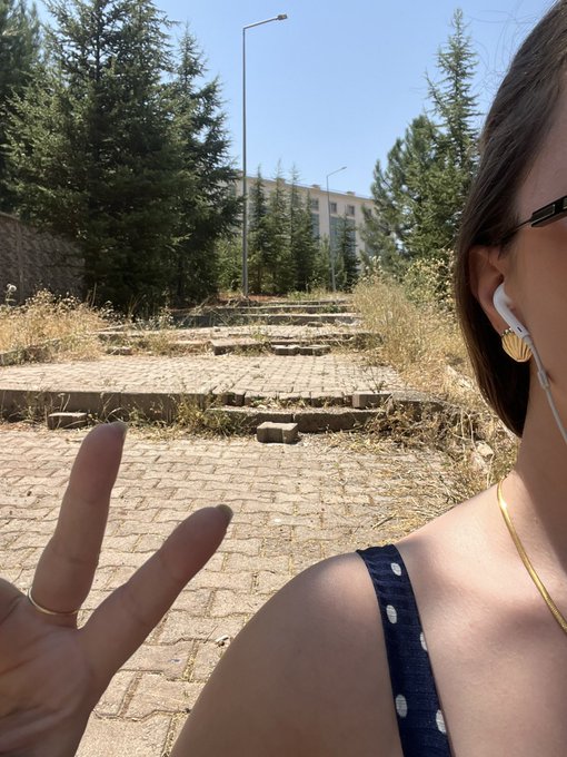 Selfie of a person wearing white wired earbuds and a dark polka-dot top making a peace sign, standing in what appears to be an abandoned or overgrown outdoor area with a building in the background.