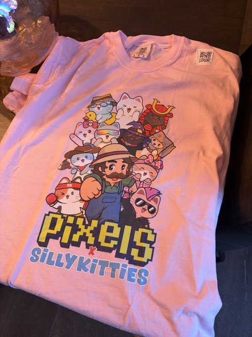 A white t-shirt lies on a wooden surface featuring cartoon artwork and text that reads 'PIXELS x SillyKitties'.