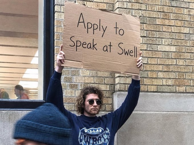 The image features a man holding up a sign that reads "Apply to Speak at Swell" while standing outside against a brick wall. The text on the sign suggests that he wants people to apply for jobs or opportunities related to the concept of "Swell", which could be an online platform, a community event, or any other gathering focused on discussing and sharing ideas about finance and its future.