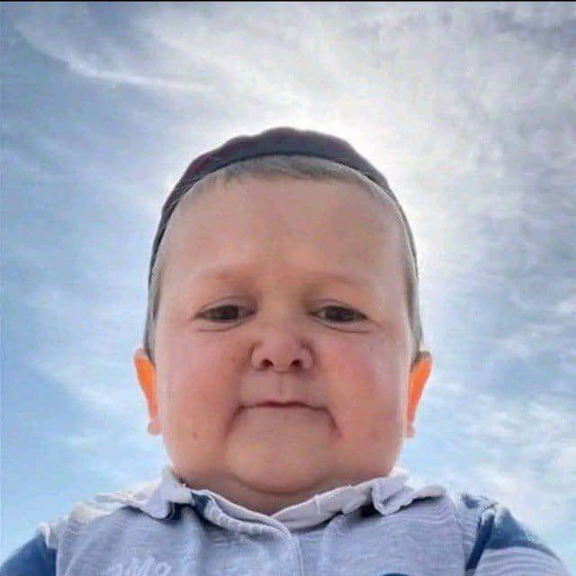 Close-up portrait of a smiling baby wearing a blue striped outfit and a small cap against a cloudy sky background.