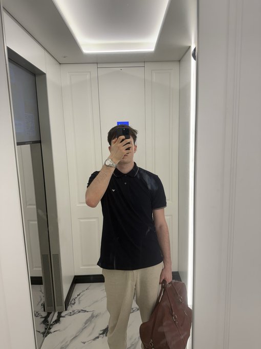 A mirror selfie of a man wearing a black polo shirt and beige pants, holding a smartphone and a brown leather bag in a white-walled room with marble flooring and an elevator.