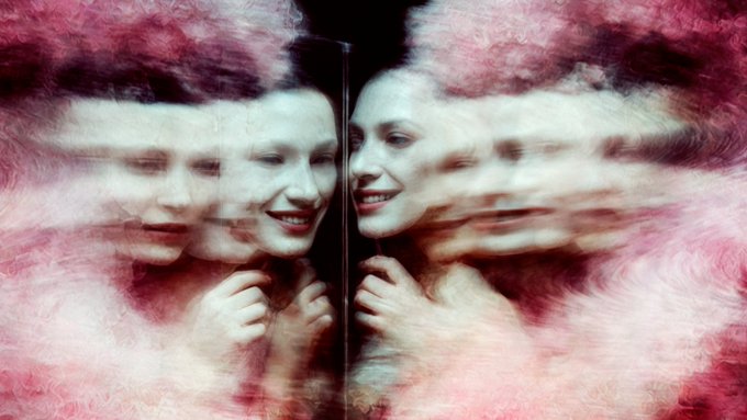 An abstract, motion-blurred image showing a smiling woman's face reflected or distorted alongside other faces in pink and white tones.