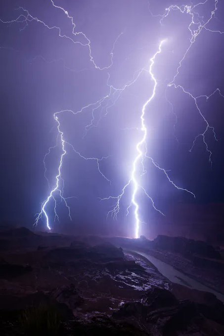 Spectacular, branching bolts of lightning illuminate a dark purple stormy sky, striking down toward a canyon landscape with a winding river far below.