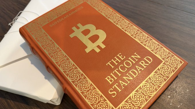 A leather-bound book titled 'THE BITCOIN STANDARD' by Saifedean Ammous sits on a wooden surface, featuring a gold Bitcoin symbol on its cover.