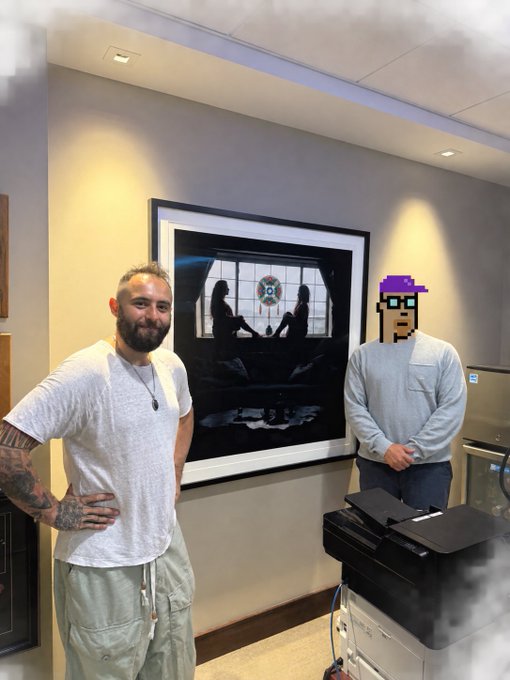 Two men stand in a room next to a framed pixel art print, with a printer in the foreground and one man's face covered by a pixel art edit.