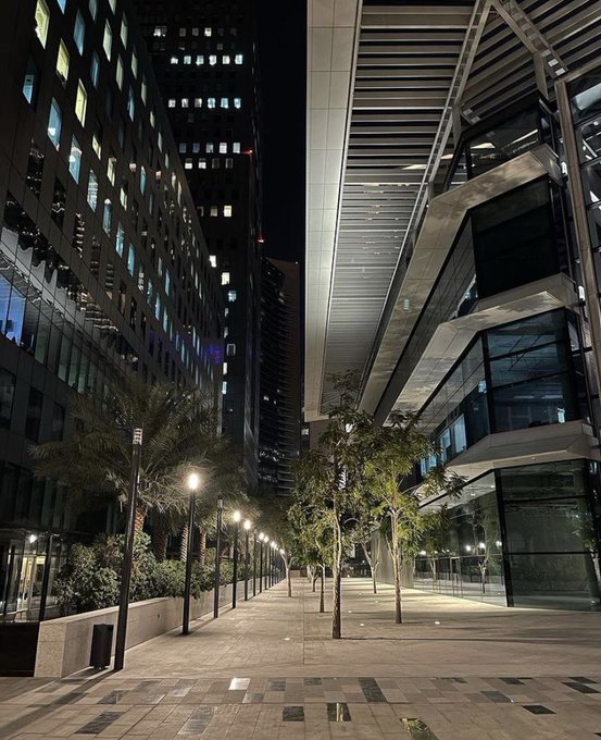 The image shows a city street at night with tall buildings lining both sides, including a modern glass building that stands out among them. The street is lined with trees, adding a touch of nature to the urban landscape. In front of one of these trees, there are two street lamps and a bench for people to sit on. A post on social media has been shared about this scene, possibly promoting an event or a new wallet that was built by the community itself.