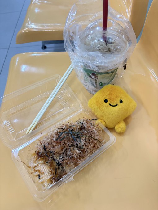 A takeout meal in a plastic container with chopsticks, accompanied by a drink in a plastic bag and a small yellow plush toy with a smiley face on an orange table.
