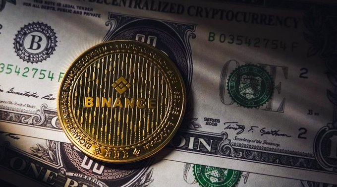 A gold Binance coin resting on top of US one-dollar bills, with the text 'DECENTRALIZED CRYPTOCURRENCY' visible on the bills.