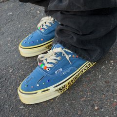 A close-up of blue and yellow sneakers covered in various charms, worn with black pants on a paved surface.