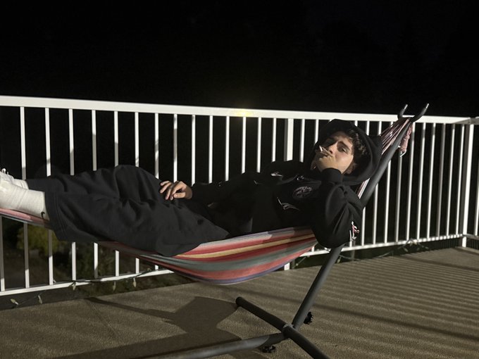 A person relaxes in a striped hammock on a deck at night, leaning back with their legs extended.