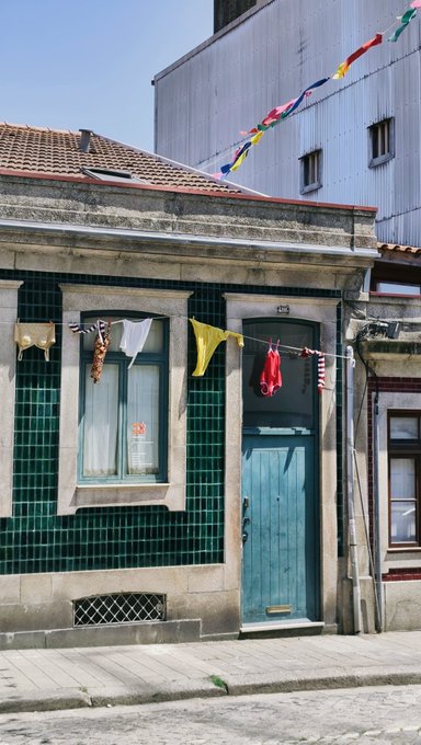 A charming traditional house in Porto, Portugal, features a vibrant green-tiled façade with clothes hanging from the window, including a yellow shirt and white garment, while colorful triangular bunting stretches overhead, suggesting a relaxed Sunday atmosphere typical of the city's festive streetscapes.