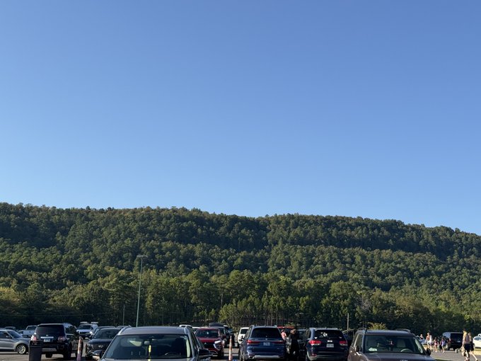 A parking lot filled with cars in the foreground, with a forested mountain ridge rising under a clear blue sky in the background.